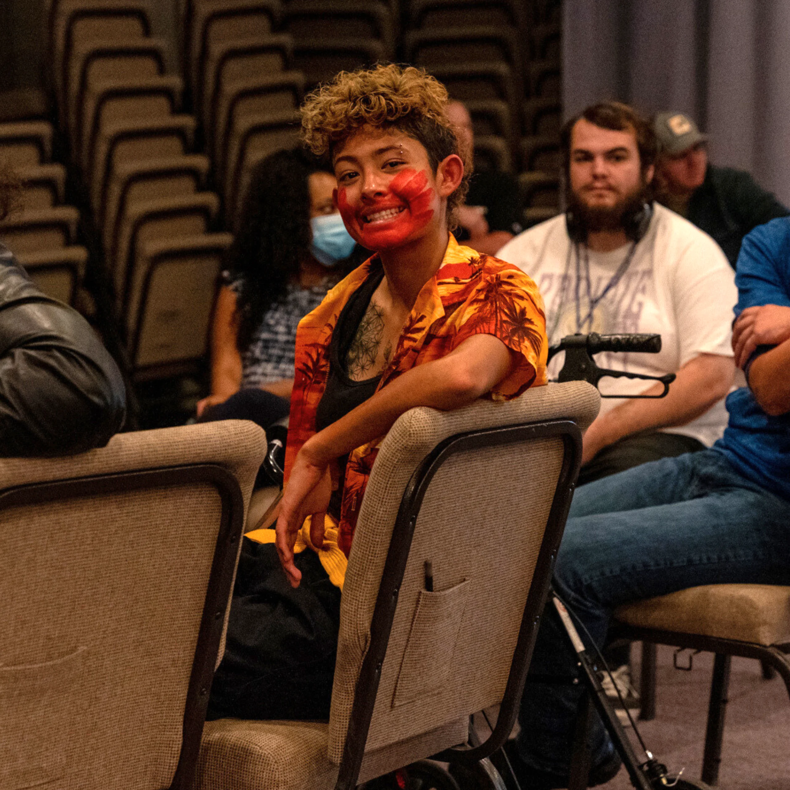 A smiling young person sits turned halfway around in a conference chair. They have a red hand print painted across their mouth and cheeks